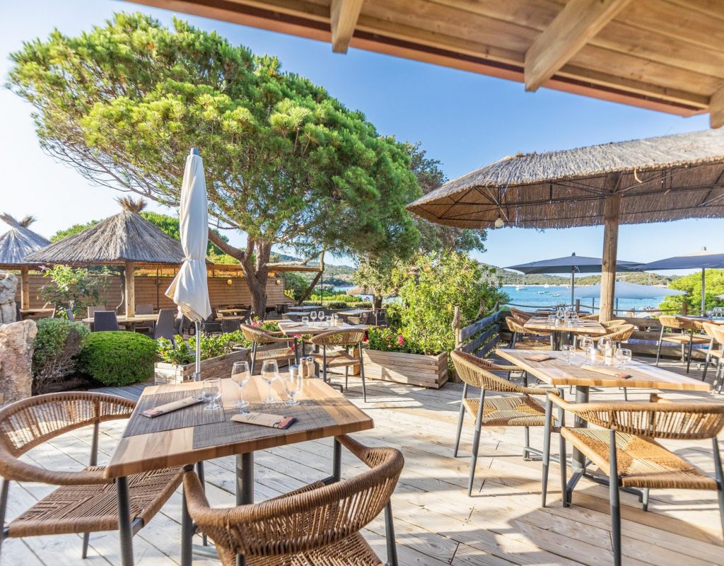 Tables dressées pour le service sur la terrasse du restaurant,  en bois et ombragée, avec vue sur la baie de Rondinara, par une journée ensoleillée
